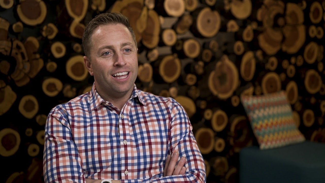 Man smiling in plaid shirt seated, decorative wood background