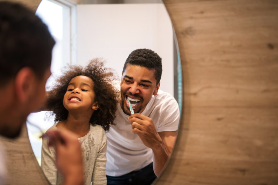 Father and daughter brushing teeth together in mirror.